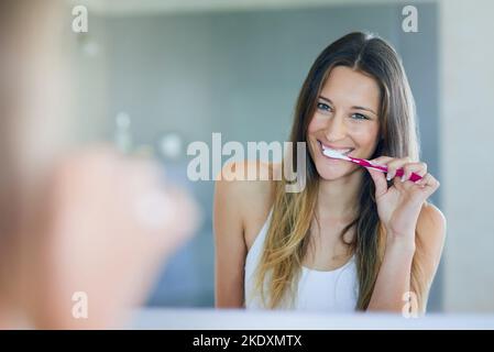 All it takes is two minutes twice a day. Portrait of young woman looking in the bathroom mirror while brushing her teeth. Stock Photo