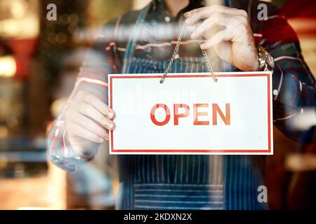 Its opening day. Closeup shot of an unrecognizable man hanging up an open sign in a shop window. Stock Photo