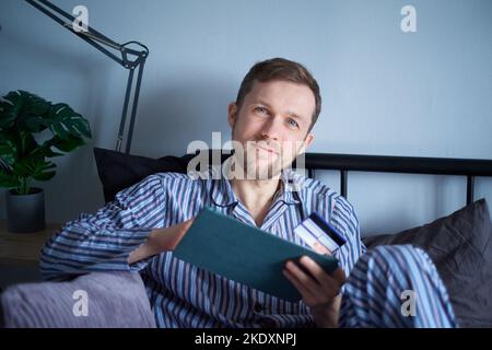 Black friday shopping concept. Young attractive man using tablet computer sitting in bed in the morning for making purchases online. Caucasian male using tablet for shopping with credit card in hands Stock Photo