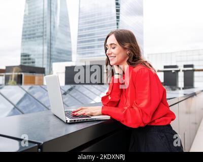 Happy woman in stylish red shirt touching neck and smiling while browsing data on netbook during work on remote project on city street Stock Photo