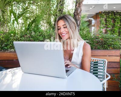 Happy blond woman smiling and browsing netbook while sitting at table and working remotely on terrace Stock Photo