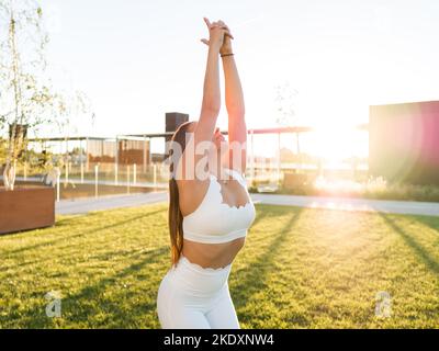 Fit young sportswoman with praying hands practicing yoga on green lawn on sunny day in city park Stock Photo