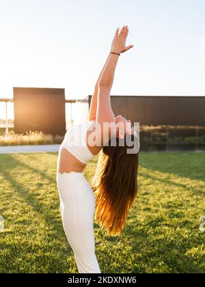 Fit young sportswoman with praying hands practicing yoga on green lawn on sunny day in city park Stock Photo