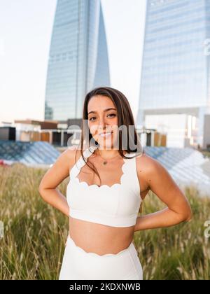 Positive young woman with long brown hair in white top and pants standing on meadow with hands behind back against modern buildings with glass walls w Stock Photo