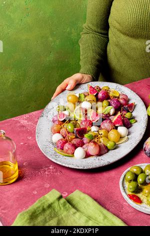 Cropped unrecognizable cook holding plate with fresh ripe grapes, olives, figs and mozzarella seasonal christmas salad placed on plate on green tablet Stock Photo