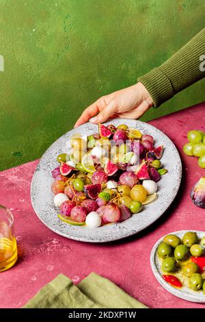 Cropped unrecognizable cook holding plate with fresh ripe grapes, olives, figs and mozzarella seasonal christmas salad placed on plate on green tablet Stock Photo