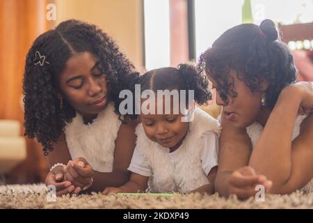 Focused Indian sisters with curly hair watching interesting video on cellphone while lying together on floor in light living room Stock Photo