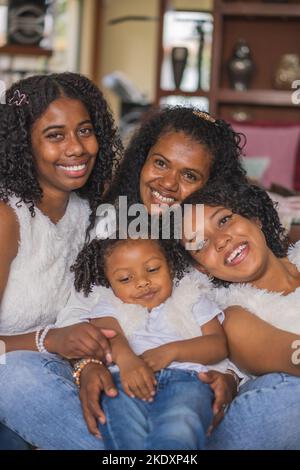 Cheerful Indian mother and daughters with curly hair in casual wear sitting together on sofa in light living room at home Stock Photo