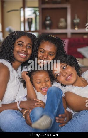 Cheerful Indian mother and daughters with curly hair in casual wear sitting together on sofa in light living room at home Stock Photo