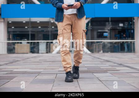 Cropped unrecognizable male in casual clothes texting on mobile phone while standing on modern city street in daytime Stock Photo