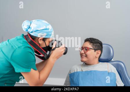 Side view of young ethnic female dentist in uniform and mask taking photo of teeth of preteen patient sitting on chair during appointment in modern de Stock Photo