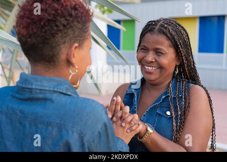Back view of unrecognizable adult black daughter in denim shirt with short hair holding hands of happy mature mom looking at camera while standing nea Stock Photo