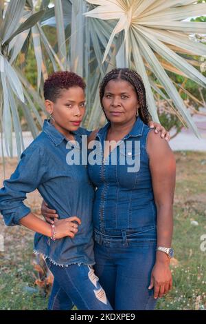 Adult black daughter in denim shirt with short hair embracing mature mom and looking at camera while standing near leaves of tropical palm in park Stock Photo