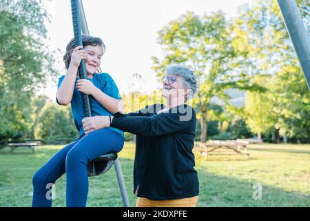 Grandma in casual clothes with glasses pushing swing with laughing granddaughter while spending sunny weekend day in summer park Stock Photo