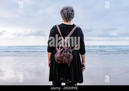 From behind elderly female traveler standing on wet sand near waving sea on gray weekend day on resort Stock Photo