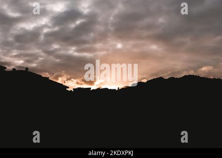 Picturesque view of bright sun setting on cloudy sky over silhouettes of houses and mountains Stock Photo