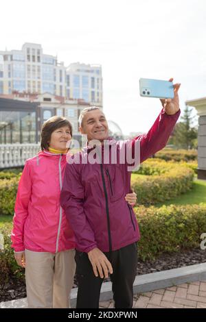 Mature couple hugging and taking selfie during break in fitness training on sunny day in park Stock Photo