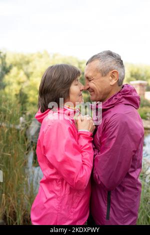 Side view of middle aged man and woman clasping hands and touching foreheads while standing near pond during romantic date in park Stock Photo