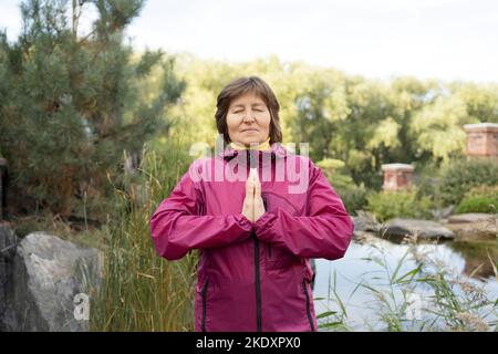 Tranquil middle aged female athlete in activewear standing with eyes closed with prayer hands meditating near small pond during fitness workout in par Stock Photo