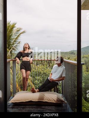 Woman leaning on fence while standing on balcony with boyfriend chilling near on chair enjoying exotic vacation in hotel Stock Photo