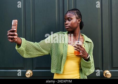 Self assured young black female millennial with dark hair in stylish outfit taking selfie on smartphone while standing near green wooden door with cup Stock Photo