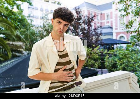 Stylish young man standing on rooftop terrace near green trees and looking at camera with hand on stomach in sunlight Stock Photo