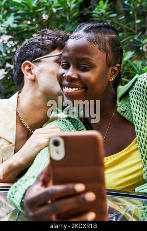 Stylish young man kissing black girlfriend while sitting at table and taking selfie via smartphone while sitting at table during romantic date in gard Stock Photo
