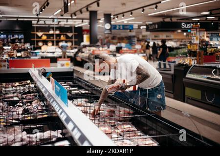 Side view of tattooed male customer in casual clothes standing near chest freezer and picking steak for dinner in supermarket Stock Photo