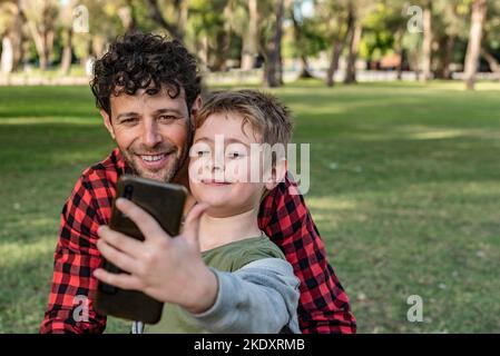 Positive son taking self portrait on smartphone together with glad dad while sitting on grass in forest in summer Stock Photo