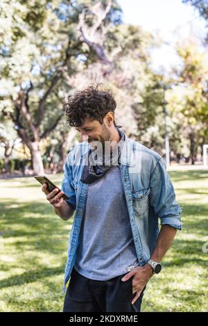 Happy male in casual clothes holding hand in pocket and browsing social media on cellphone while standing on sunlit lawn on summer weekend day in park Stock Photo