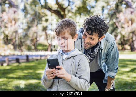 Glad dad smiling and watching son playing videogame on cellphone while spending sunny weekend day in park together Stock Photo
