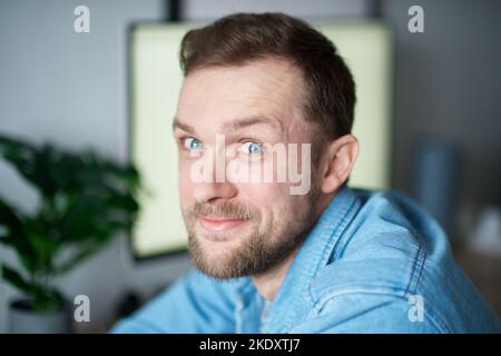 Cute bearded portrait of caucasian male freelance software engineer sitting at working place in office looking at camera with computer on background. Young handsome developer man in denim shirt Stock Photo