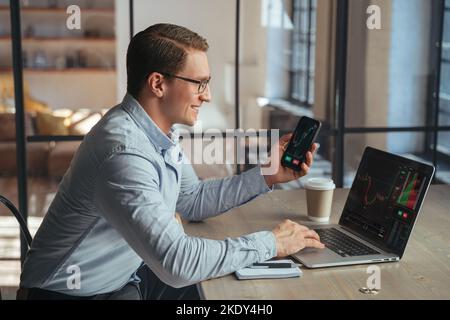 Smiling businessman crypto trader holding smartphone checking candlestick graph on laptop, buying and selling currency, happy to notice tendency of btc rising up, golden bitcoin near on table Stock Photo