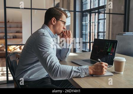 Serious crypto trader looking at laptop screen with chart, analyzing risks of buying cryptocurrency in the conditions of changing world and global recession, sitting at his office table, making notes Stock Photo
