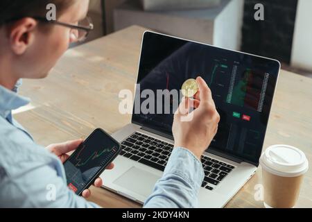 Closeup shot of successful man crypto trader or broker holding gold bitcoin, sitting at office table with laptop, investing in btc, using cryptocurrency app Stock Photo
