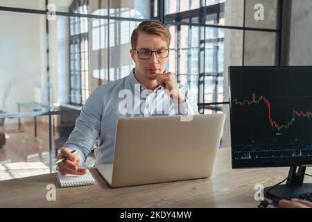 Male crypto trader working on his laptop at co-working office, looking at screen with concentrated and curious face, watching tutorial video of digital money investments and trading strategy Stock Photo