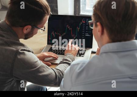 Over shoulder view of trader couch teaching colleague basics of cryptocurrency investments, explaining market trends or consulting his client, analyzing charts of money flow together on pc screen Stock Photo