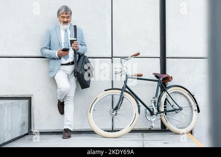 Businessman, phone and coffee with bicycle in city on social media, web or internet browsing. Tech, bike or senior manager drinking tea while on 5g Stock Photo