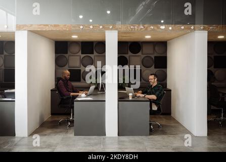 Wide angle view of two freelance business men working in cubicle styled co working office space. Stock Photo