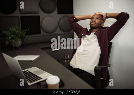 Black African man working from cubicle booth in a co working workplace. Sitting in chair looking up with hands on his head. feeling exhausted and stre Stock Photo