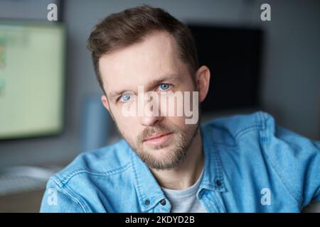 Portrait of young confident freelancer guy at home office. Cute bearded caucasian male software engineer sitting at working place looking at camera with computer on background. High quality image Stock Photo