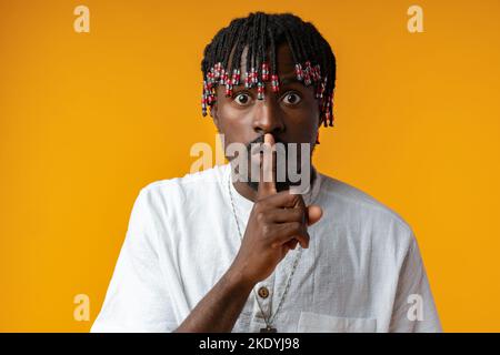 Portrait of mysterious silent african man making hush gesture over yellow background Stock Photo
