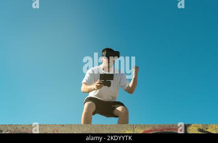 A young male trying out a VR headset doing a punching pose under a clear blue sky Stock Photo