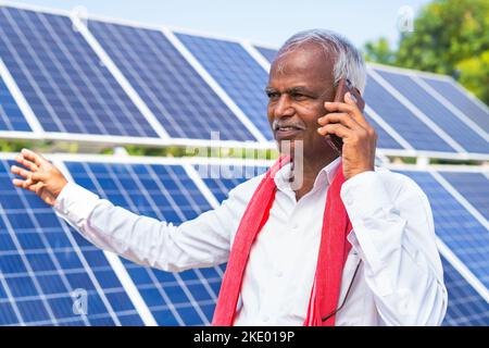 happy Indian farmer talking on mobile phone in front of solar panels about electric power generation farmland - concept of technology and modern Stock Photo