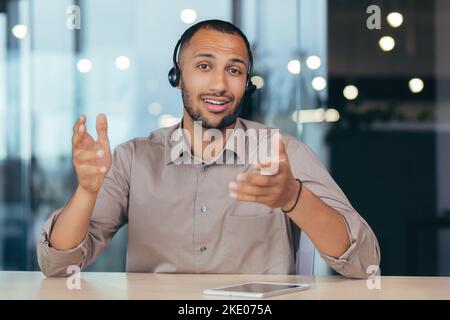 Webcam view, happy african american man talking online video call looking at camera using headset, man working inside modern office building tech support and helpline call center. Stock Photo