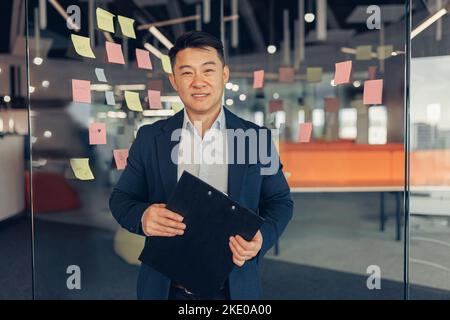 Portrait of handsome asian businessman in suit standing in office and looking at camera Stock Photo