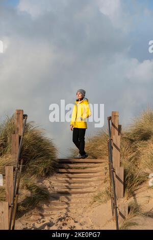 Woman wearing a yellow coat standing at top of sand dunes, looking at the view. Stock Photo