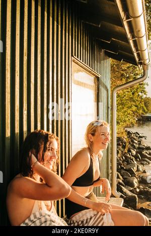 Smiling young female friends sitting by cottage during sunset Stock Photo