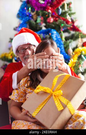 Man dressed as Santa Claus covering a little girl's eyes as she receives a gift for Christmas. Space for text. Stock Photo