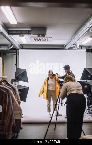 Photographer photographing designer helping model wearing jacket against white backdrop in studio Stock Photo